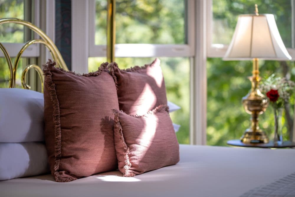 Brass bed with white linens and brown pillows next to a large wall of windows and a bedside table with a lamp and vase of flowers